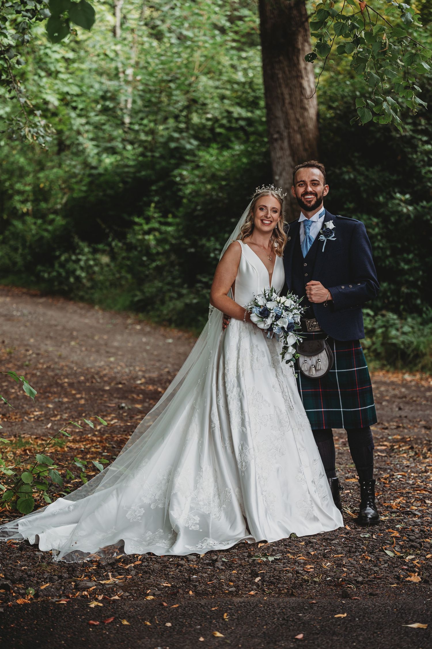A wedding couple poses on a forest path with the groom wearing a traditional Scottish kilt and the bride in a white gown.
