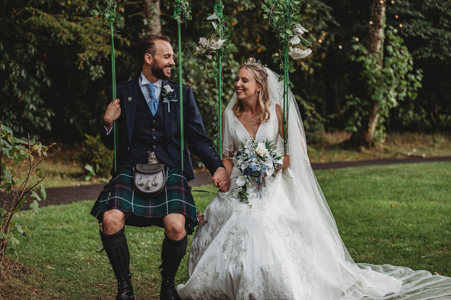 A bride in white dress and groom in traditional Scottish kilt walk together while holding hands in a lush garden setting.