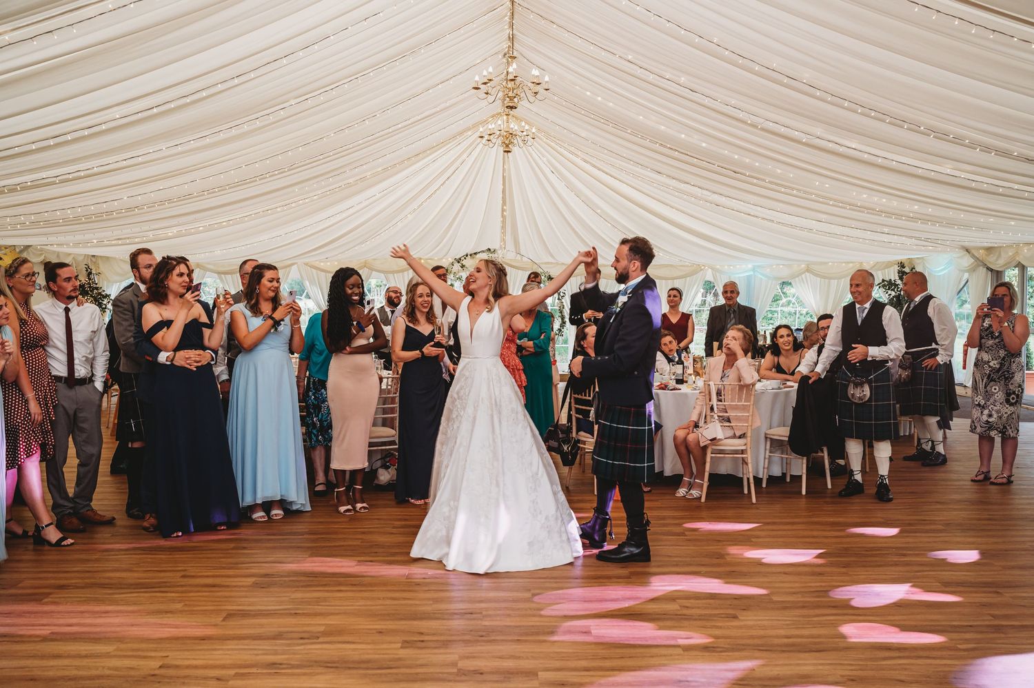 Wedding celebration in a white tent marquee with guests gathered on a wooden dance floor under elegant draping and lighting.