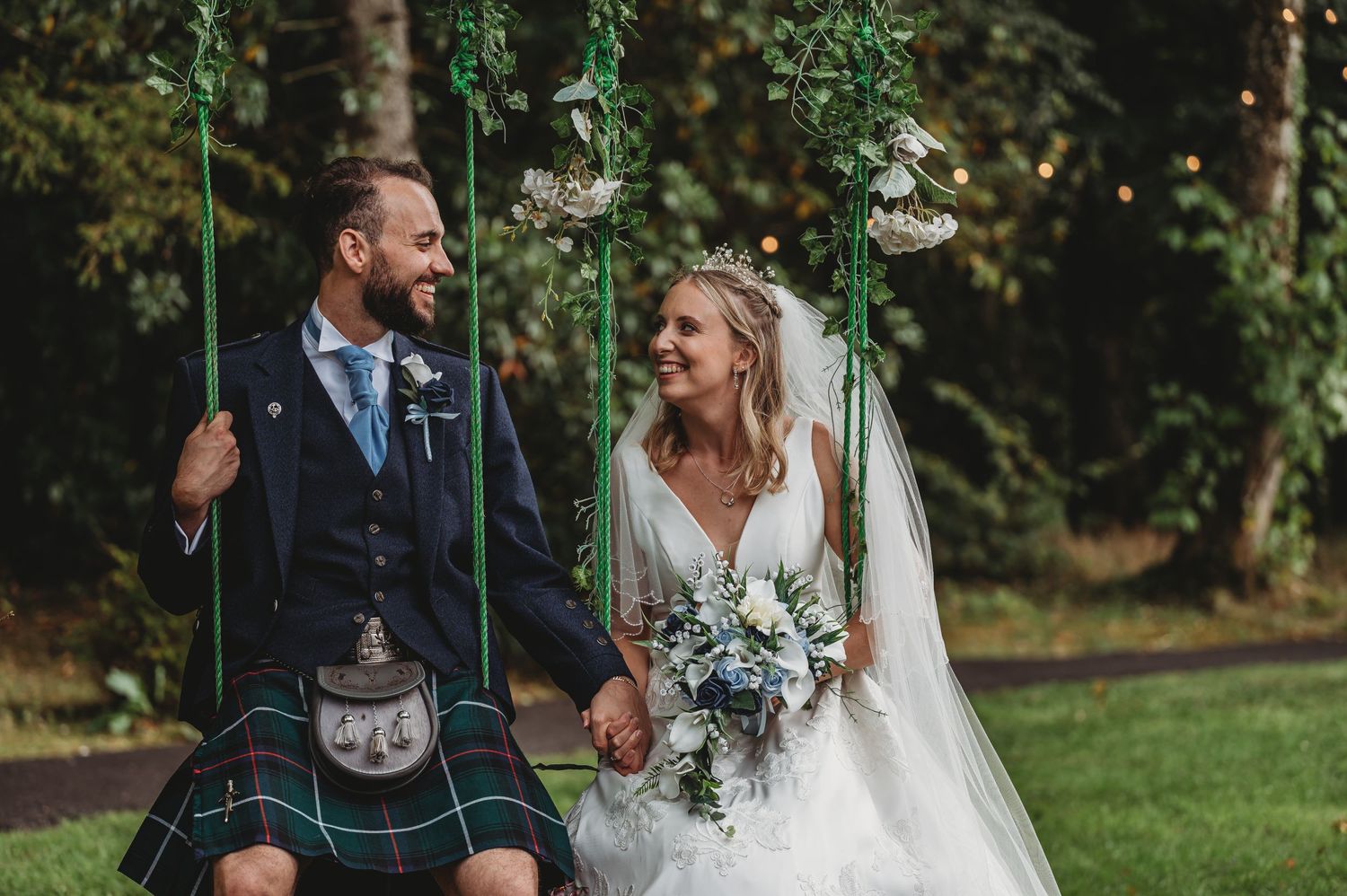 Joyful couple sits on swings during their wedding ceremony in a garden setting, with groom wearing traditional Scottish kilt.