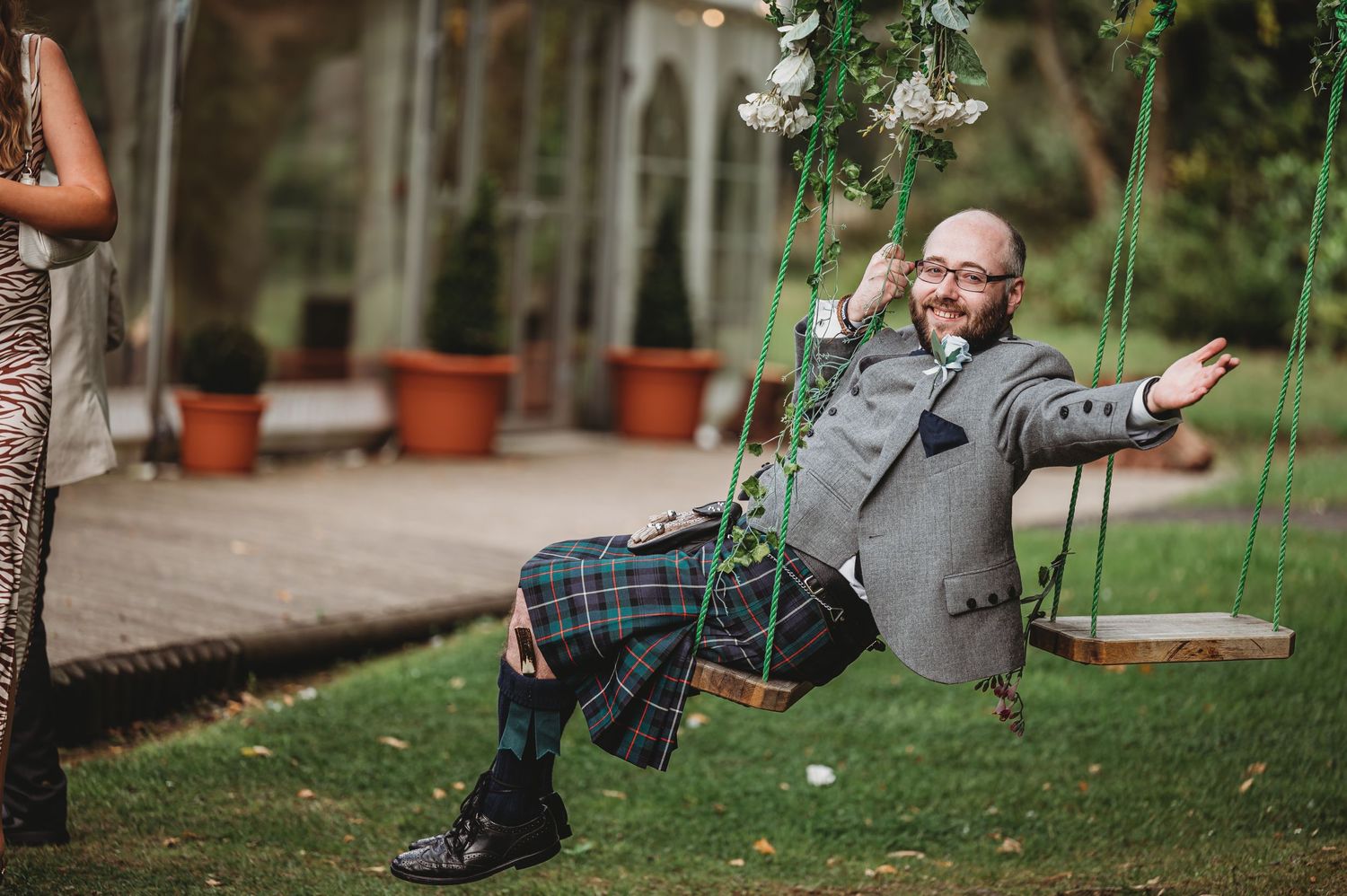 A joyful child in a gray sweater swinging outdoors near potted plants and greenery.