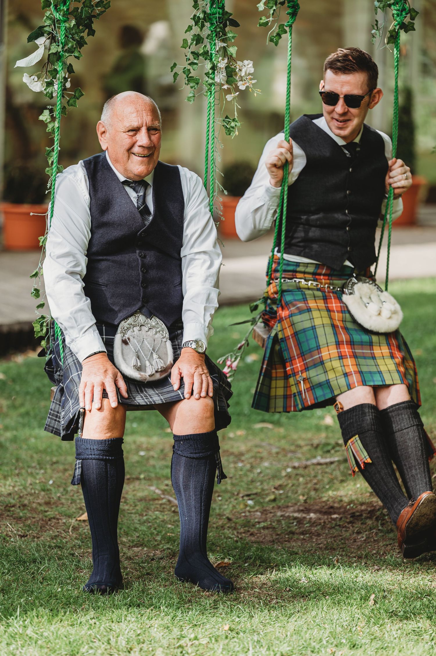 Two people wearing traditional Scottish kilts and formal attire swing together on green-decorated swings in a garden setting.