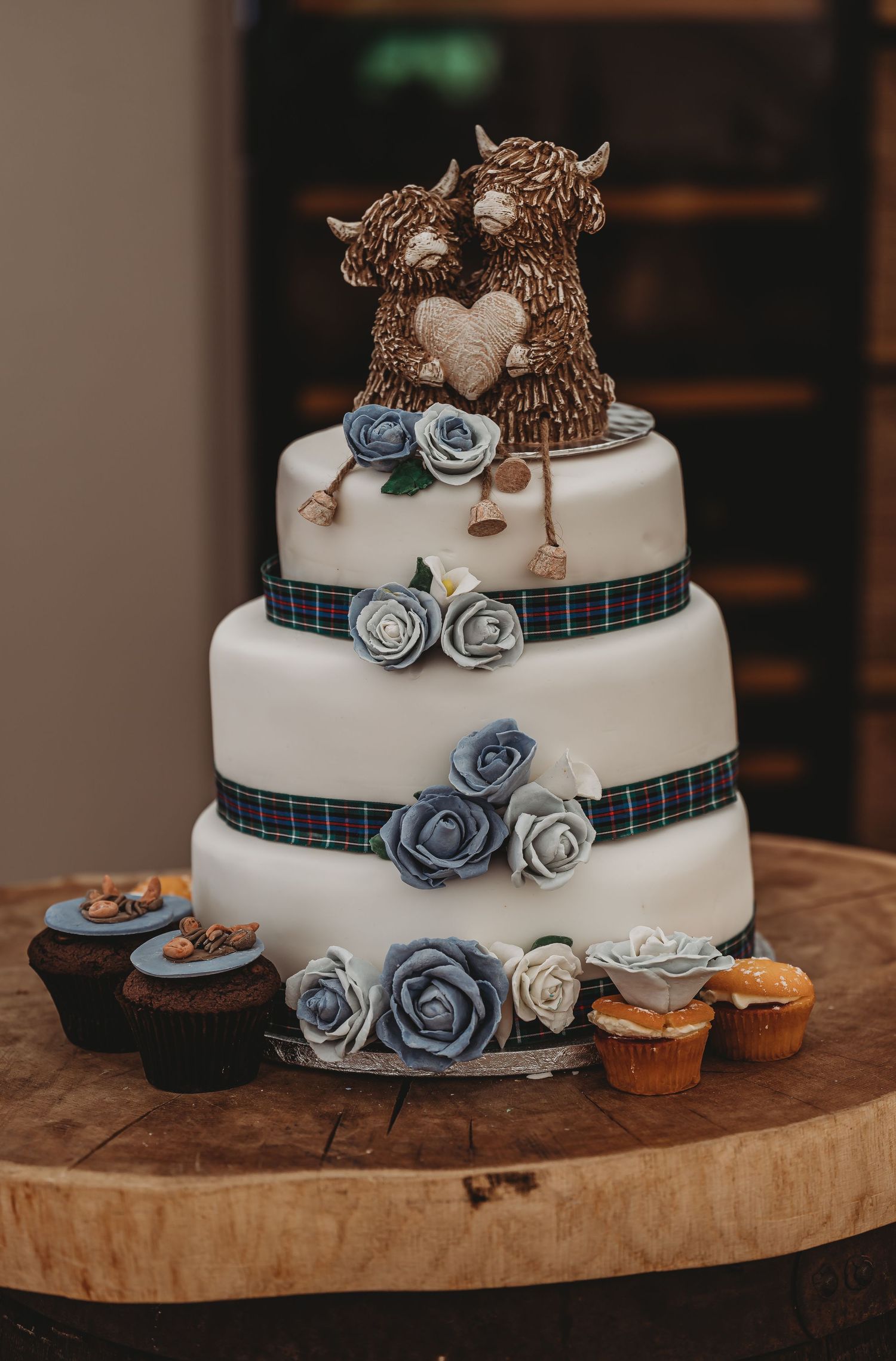 Three-tier white wedding cake with tartan ribbon, blue roses, and brown bear cake toppers displayed on a wooden slab.