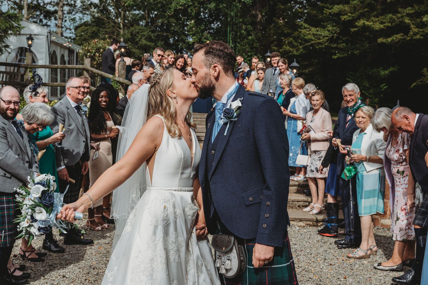 A wedding couple shares a kiss on a gravel path as guests line up to celebrate at an outdoor ceremony.