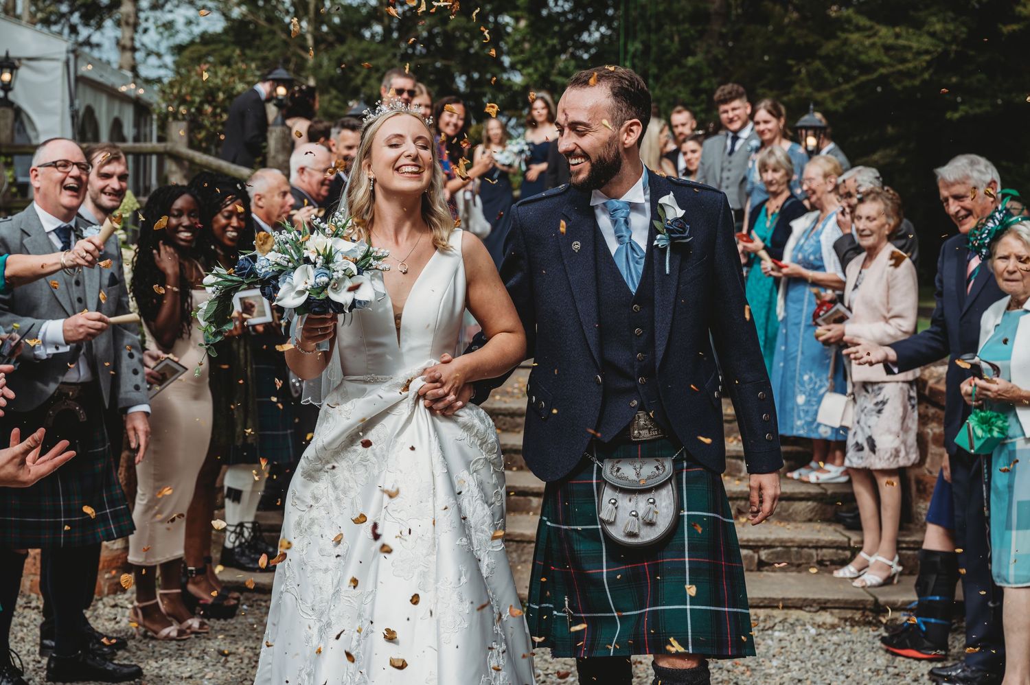 Wedding celebration with confetti being thrown as a couple in traditional Scottish attire walks through cheering guests.