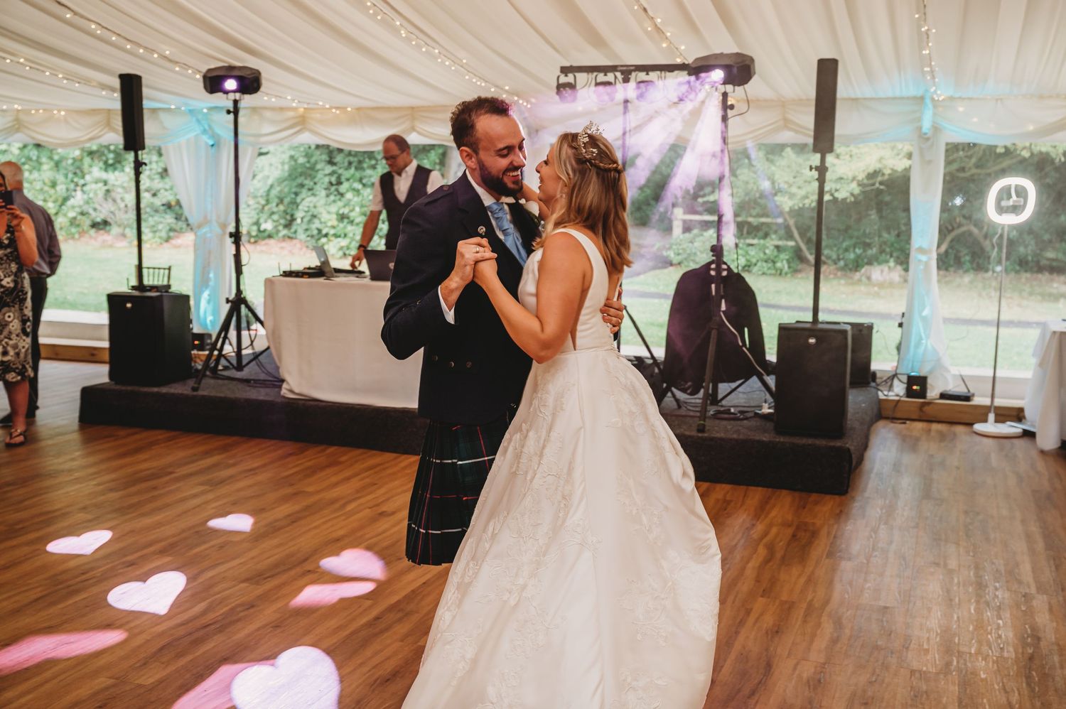 A couple shares their first dance as newlyweds on a wooden dance floor under a white tent with music equipment behind them.
