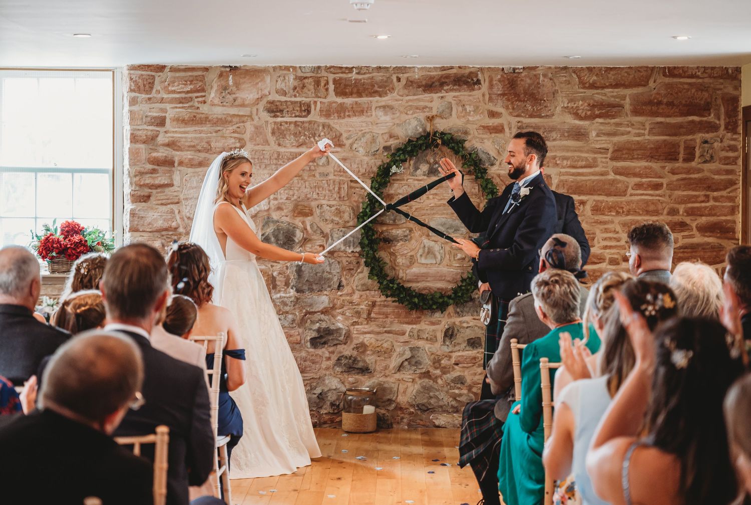 A couple performs a lightsaber duel during their wedding ceremony in front of guests and a rustic brick wall.