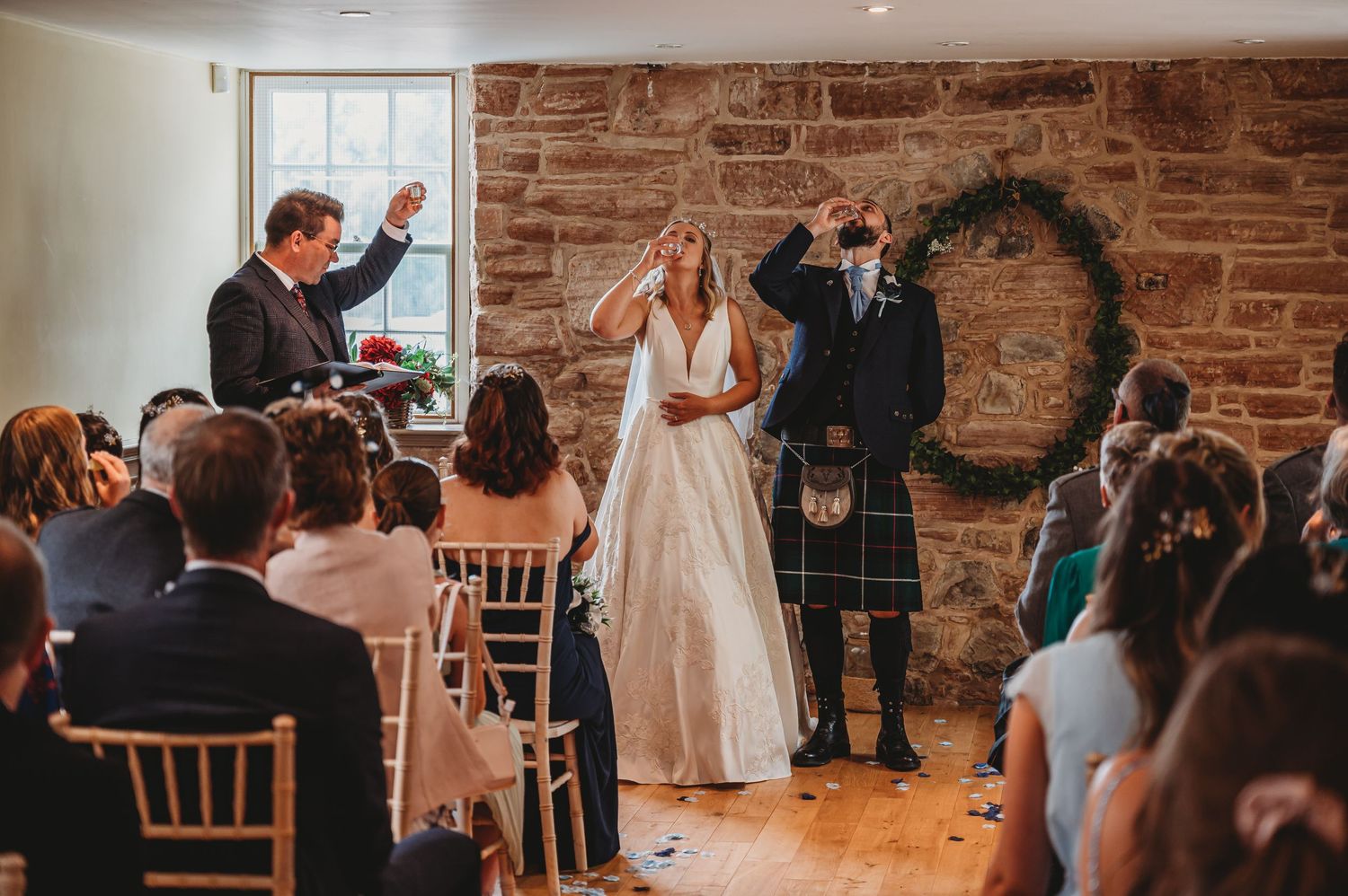 Wedding ceremony takes place in rustic brick-walled venue with guests seated in wooden chairs watching couple and officiant.