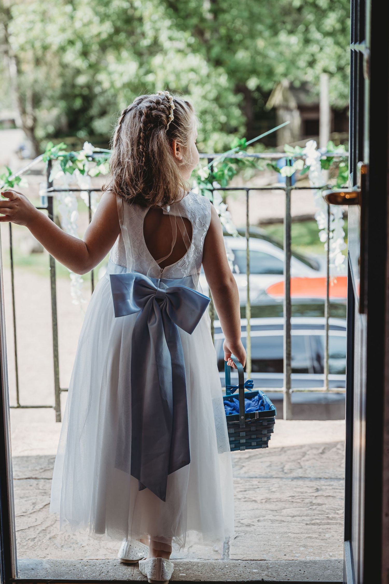 A small child in a white tulle flower girl dress with a blue bow sash stands in a doorway holding a basket.