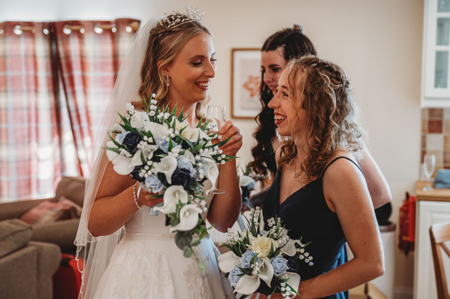 A bride and bridesmaid share a joyful candid moment while holding white and blue floral bouquets.
