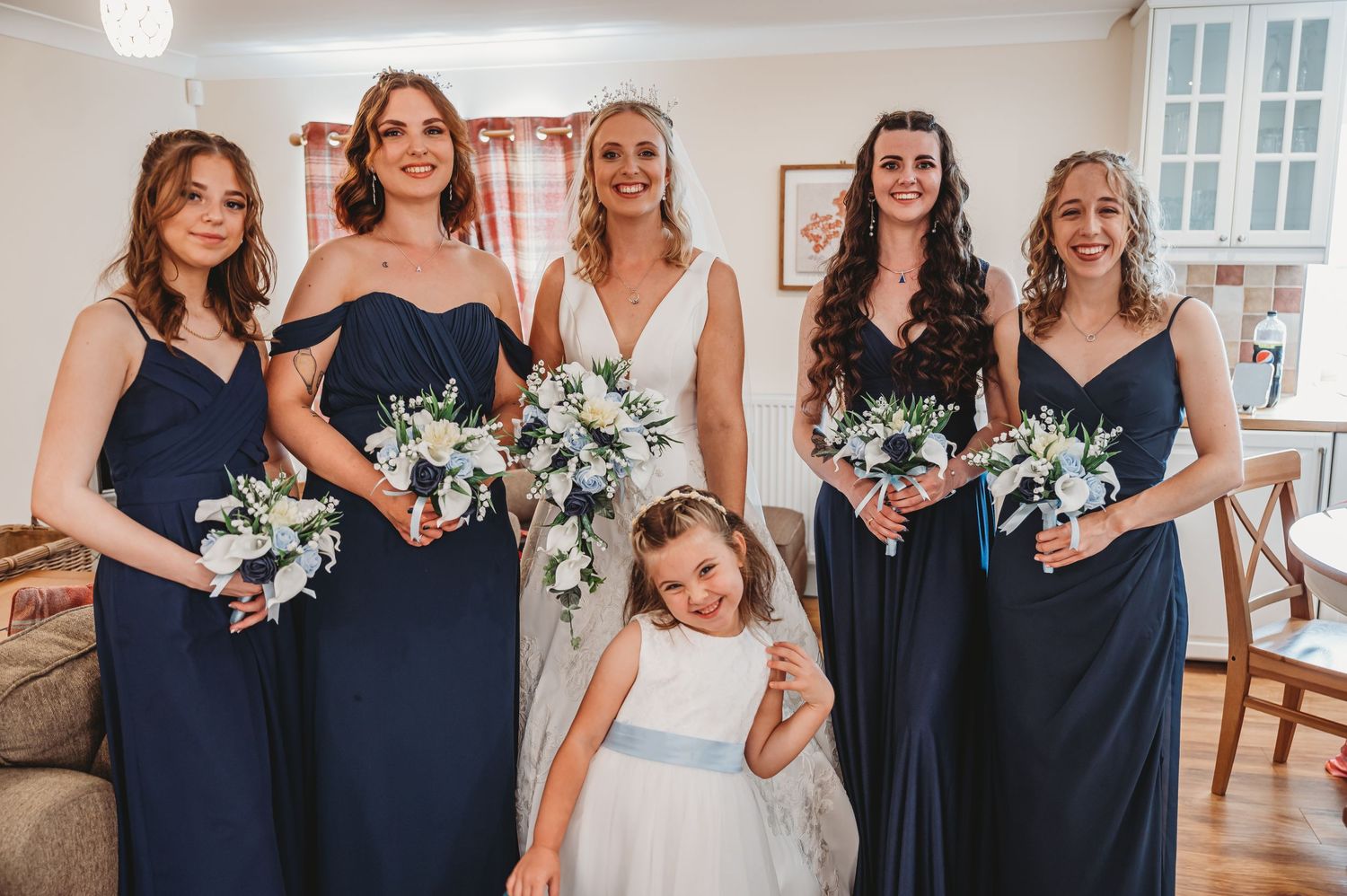 Wedding party in navy blue dresses and a flower girl in white stand together holding white bouquets indoors.