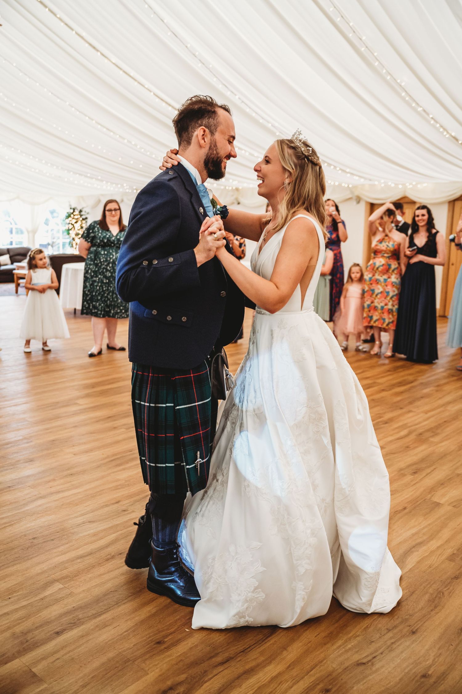 A bride in white gown dances with groom wearing traditional Scottish kilt during their wedding reception.