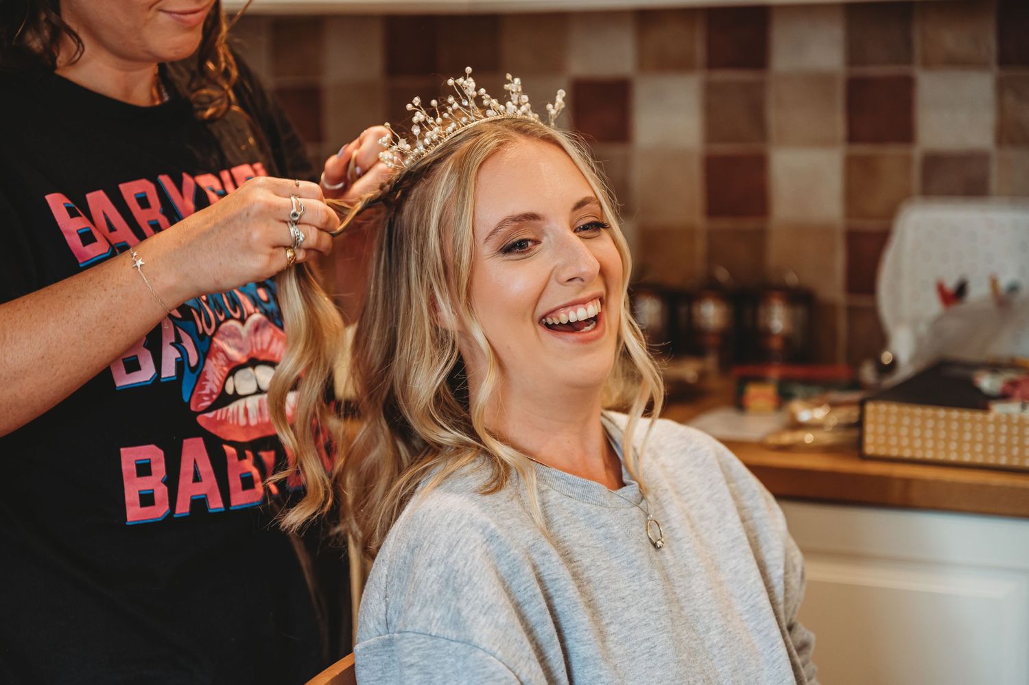 Someone laughs joyfully while getting their hair styled and having a tiara placed on their head in a kitchen setting.