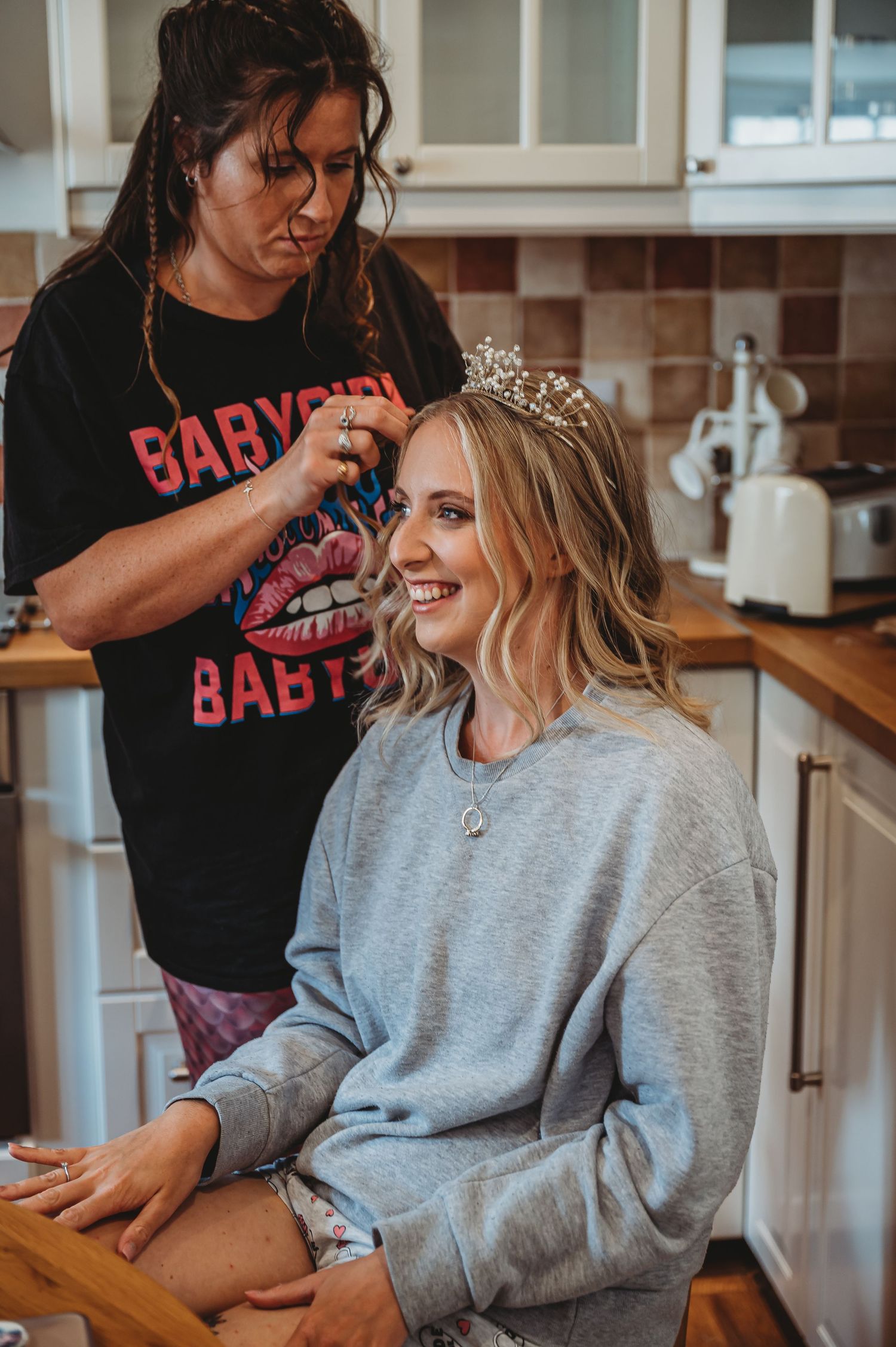 Hair stylist wearing rock band t-shirt works on bridal hair styling in modern kitchen setting.