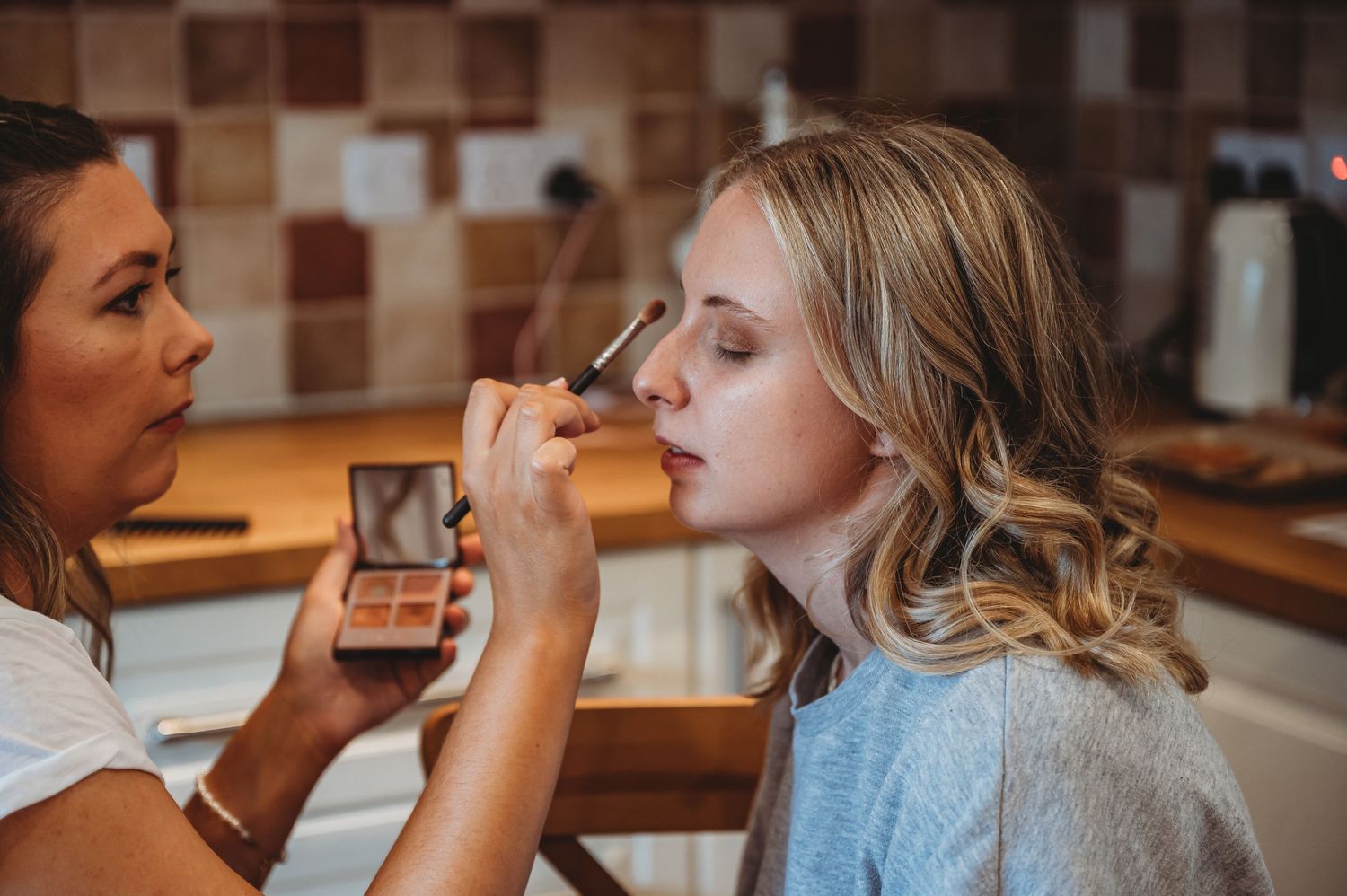 Makeup artist applying eyeshadow at vanity with brown tiled backsplash in background.