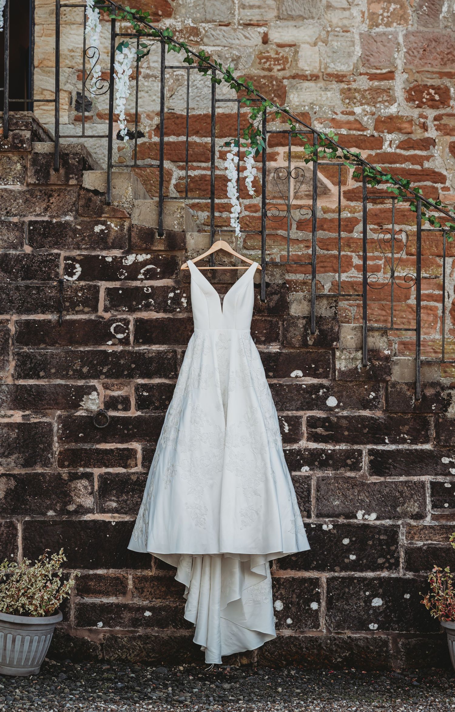 A white wedding dress hangs on a rustic brick wall with ivy-covered railing, creating an elegant bridal photo backdrop.
