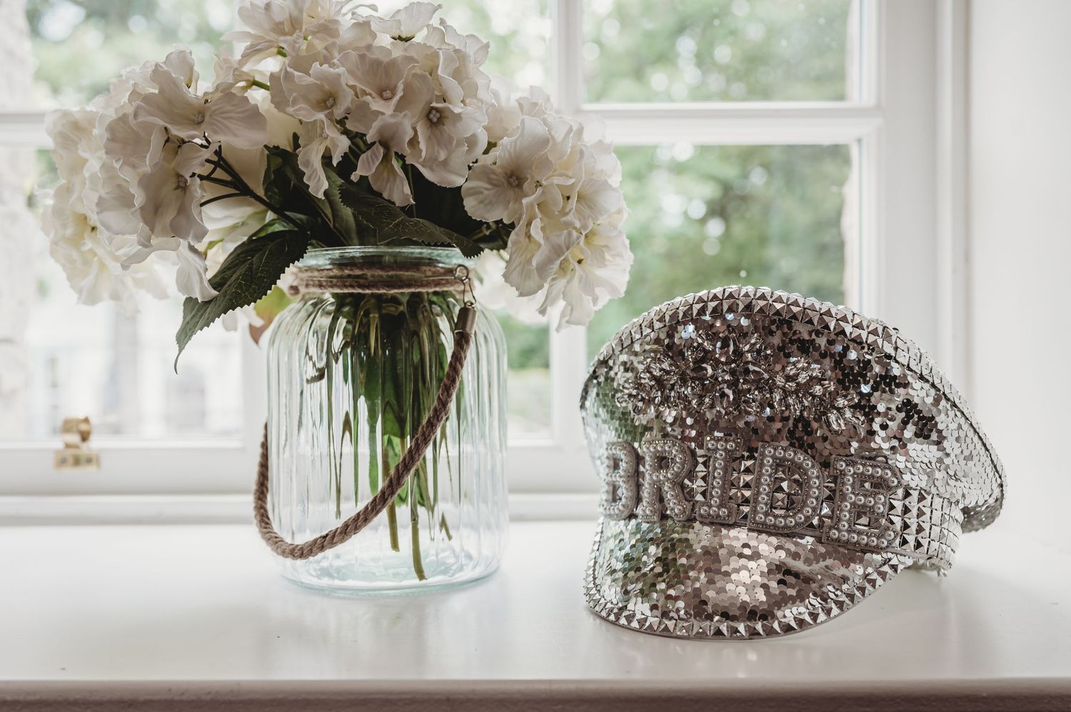 A crystal embellished baseball cap reading 'BRIDE' sits beside white hydrangeas in a mason jar near a window.