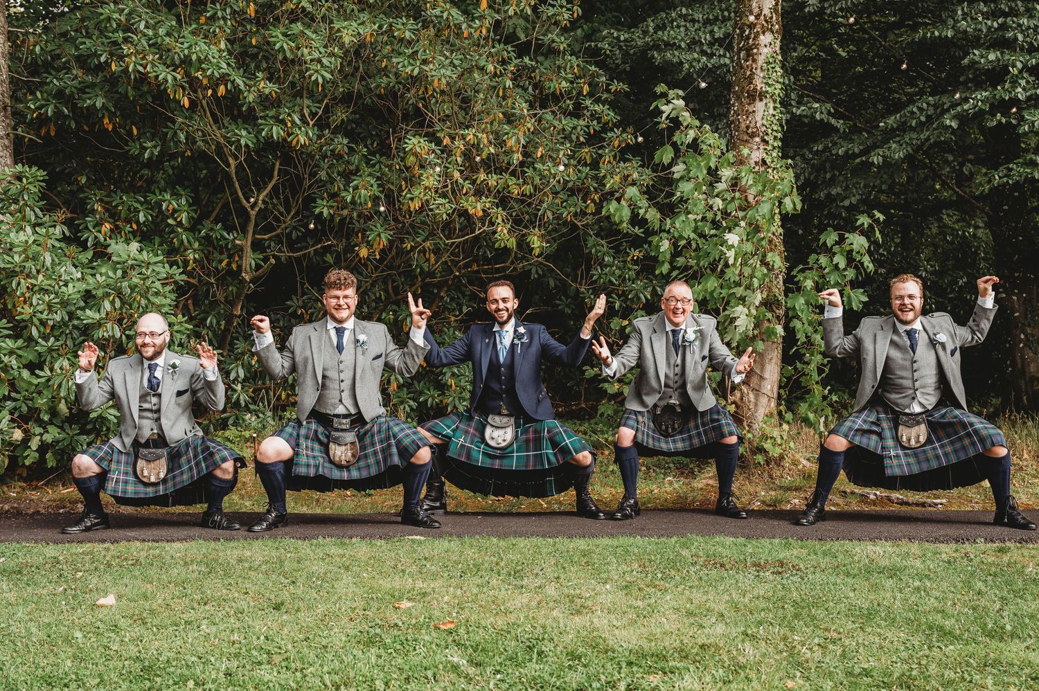 Five groomsmen in matching gray suits strike playful jumping poses together on grass at an outdoor wedding venue.