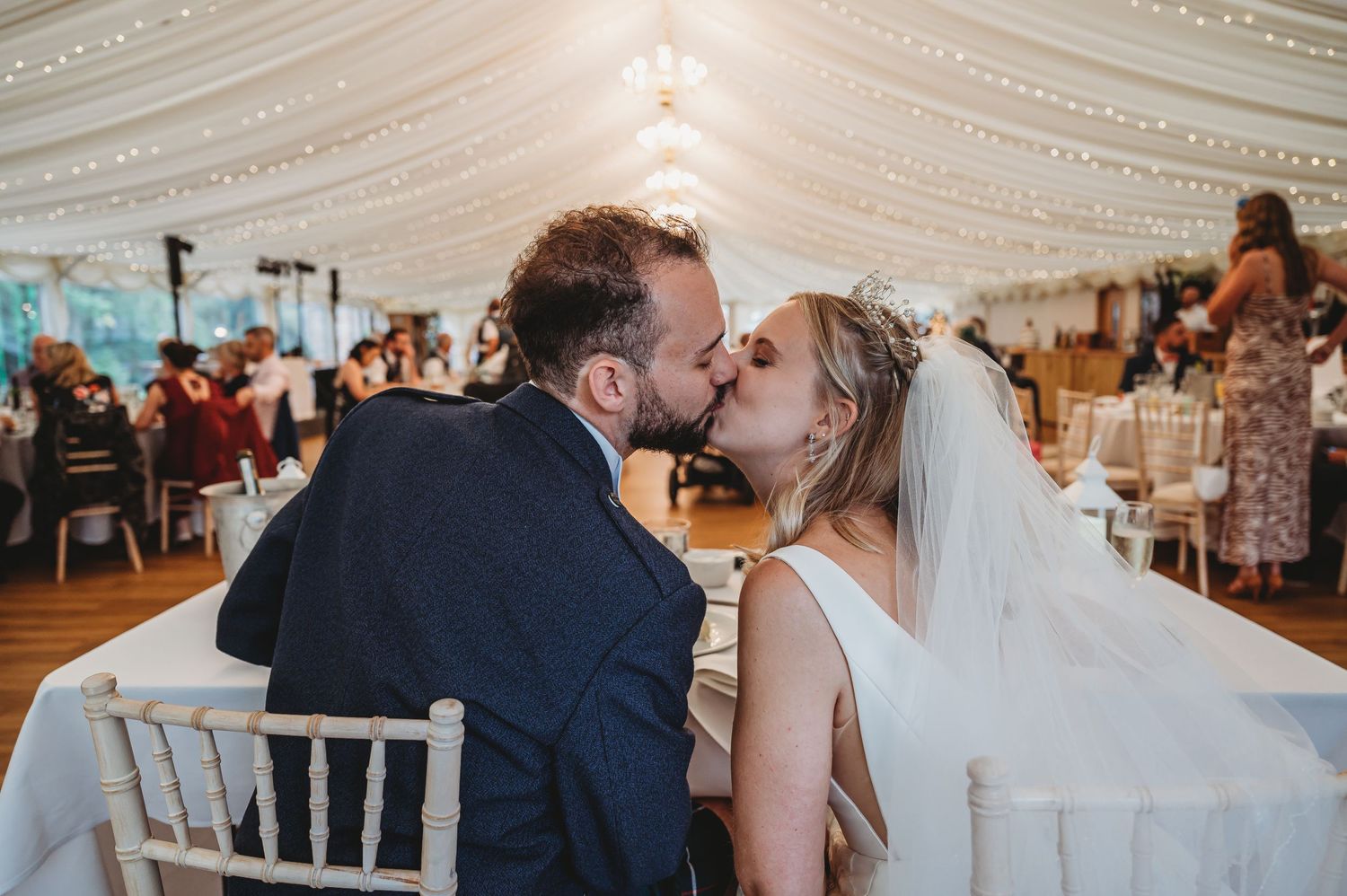 A couple shares a tender kiss during their wedding reception inside an elegant white tent decorated with chandeliers.