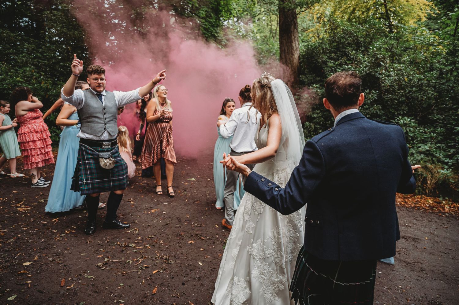 Wedding celebration in a forest with pink smoke and guests gathered on a woodland path during an outdoor ceremony.