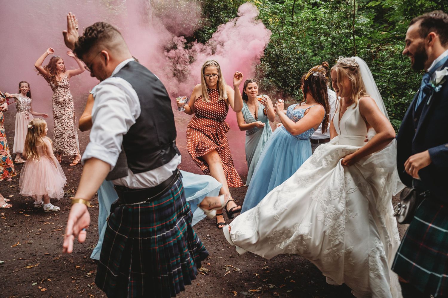 Wedding party celebrates with pink smoke bombs in a wooded setting as bride in white gown and guests in kilts dance together.