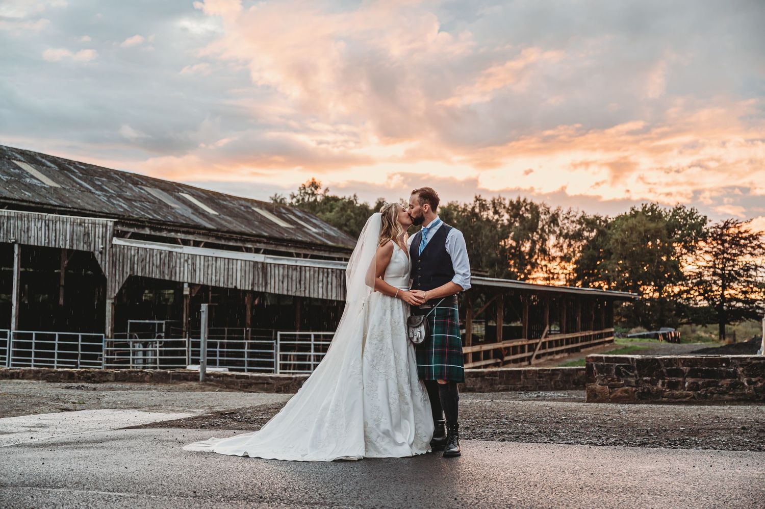 Couple in wedding attire share romantic moment at sunset outside rustic barn with glowing pink clouds in background.