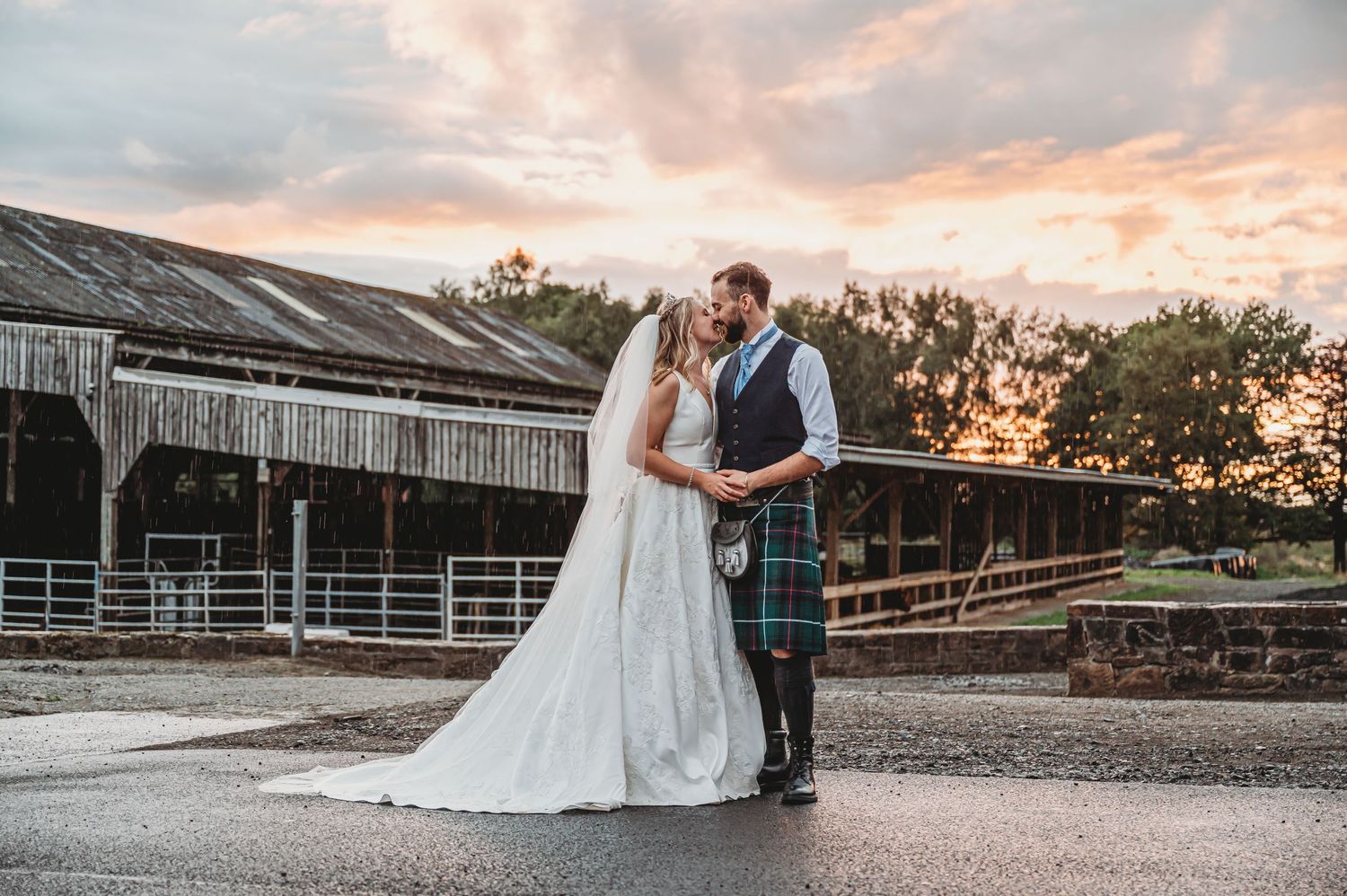 A couple in traditional Scottish wedding attire standing together at sunset by a rustic wooden barn.