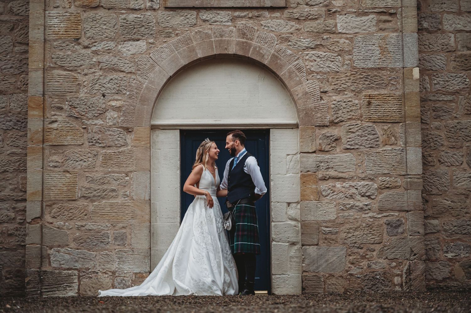 A wedding couple embraces in front of an arched stone doorway of a historic building.