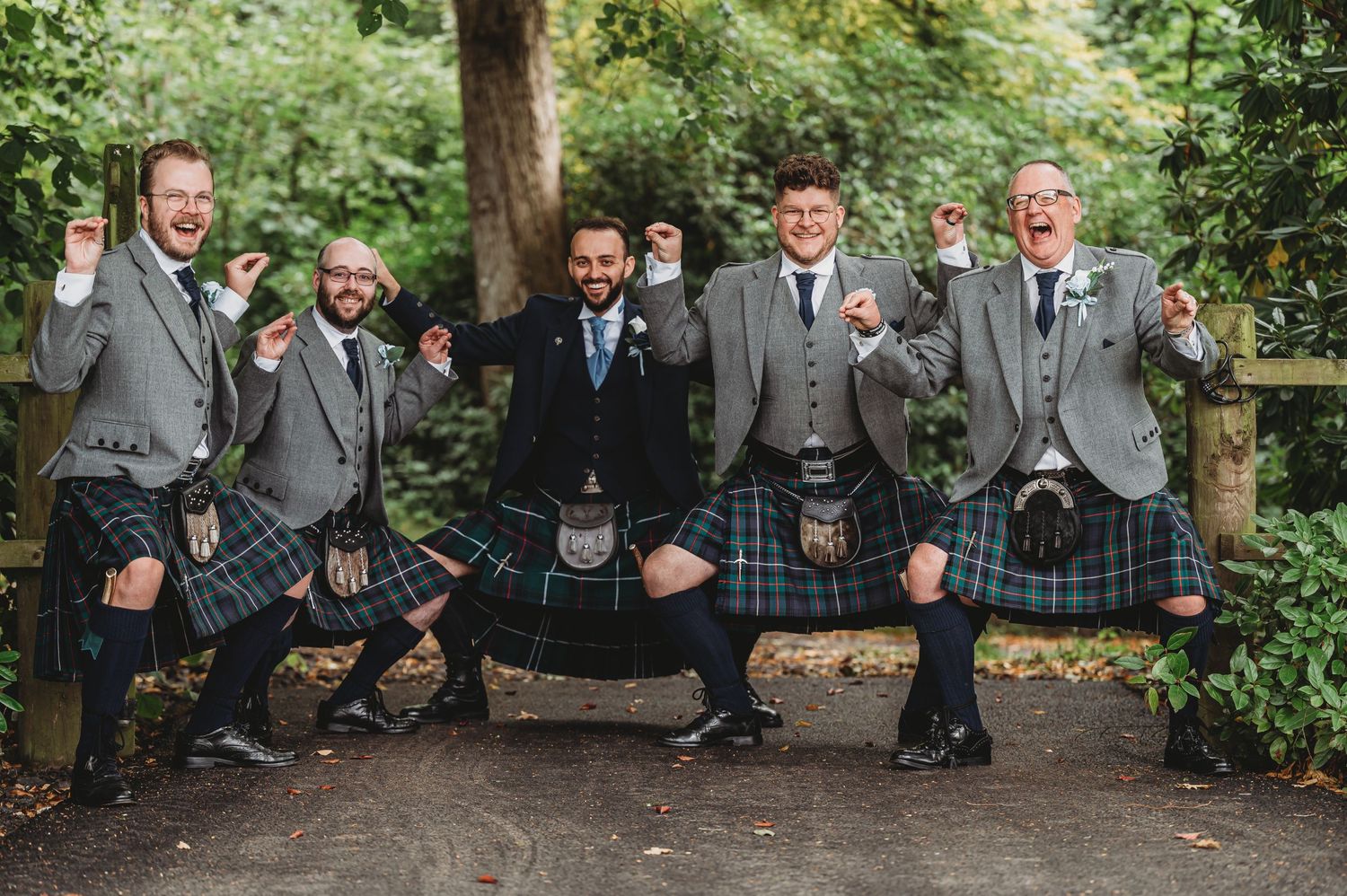 A group of groomsmen in traditional Scottish kilts and gray jackets pose playfully on a wooden bridge in a garden setting.