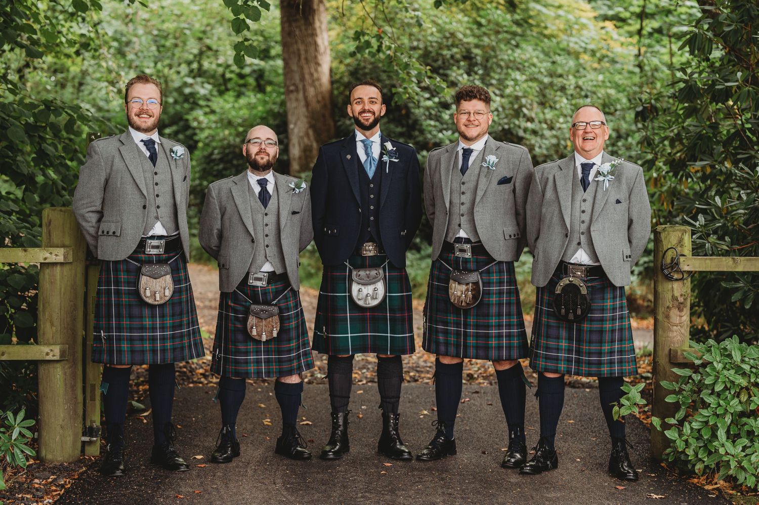 Wedding party in matching gray suits and traditional Scottish kilts pose together on woodland path at outdoor ceremony.
