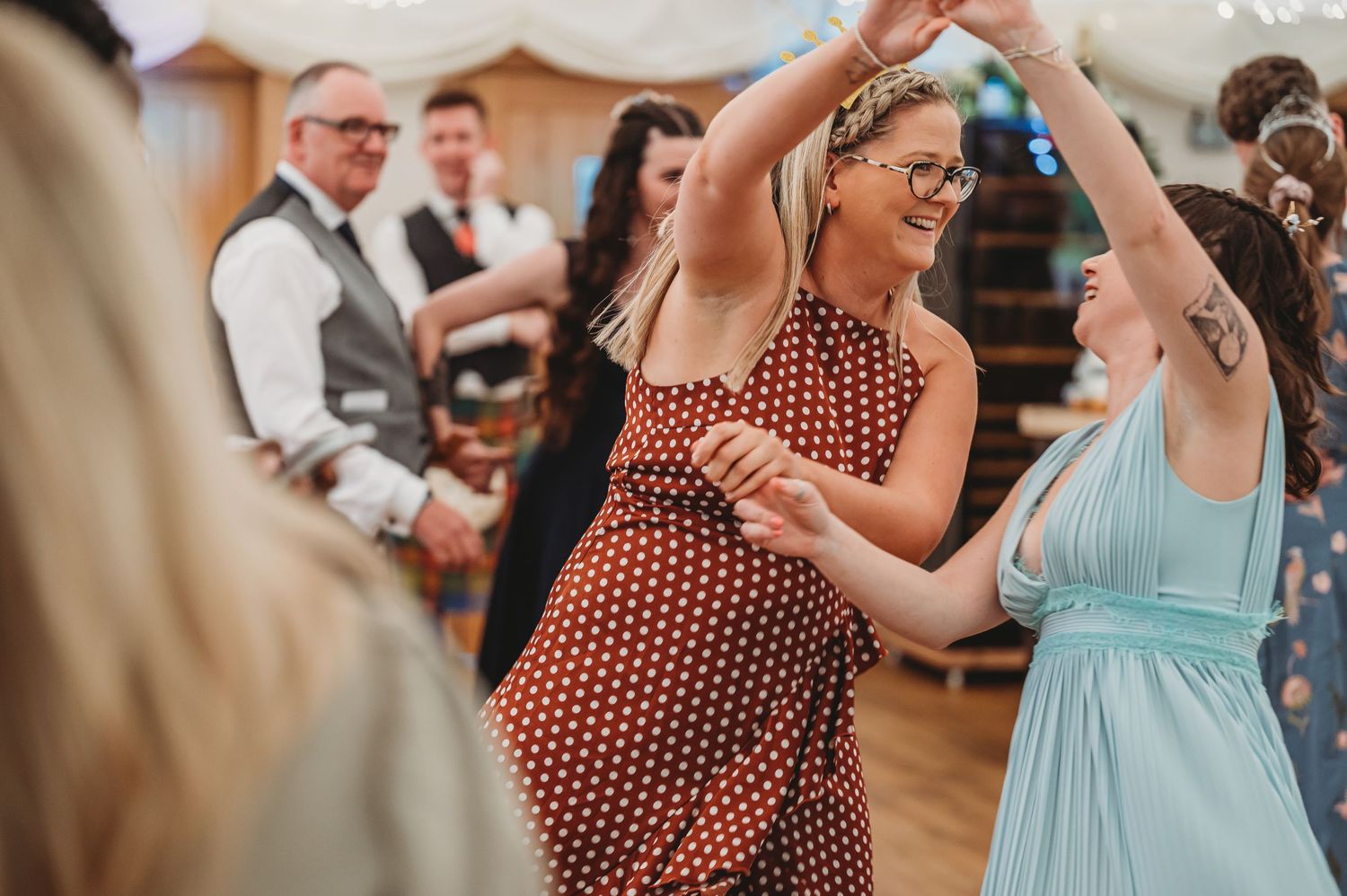 Dancing guests enjoy a lively reception in a wood-floored venue, with one wearing a polka dot dress and another in blue.