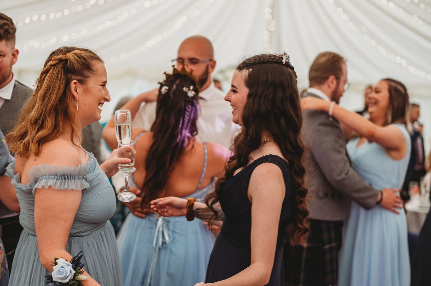 Wedding reception guests in light blue bridesmaid dresses and formal attire chatting under a white tent.