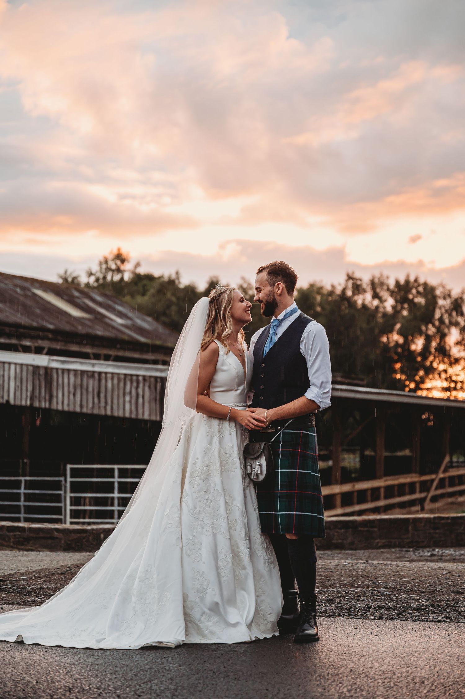 Newlywed couple embraces at sunset outside rustic barn with groom wearing traditional Scottish kilt and bride in lace gown.