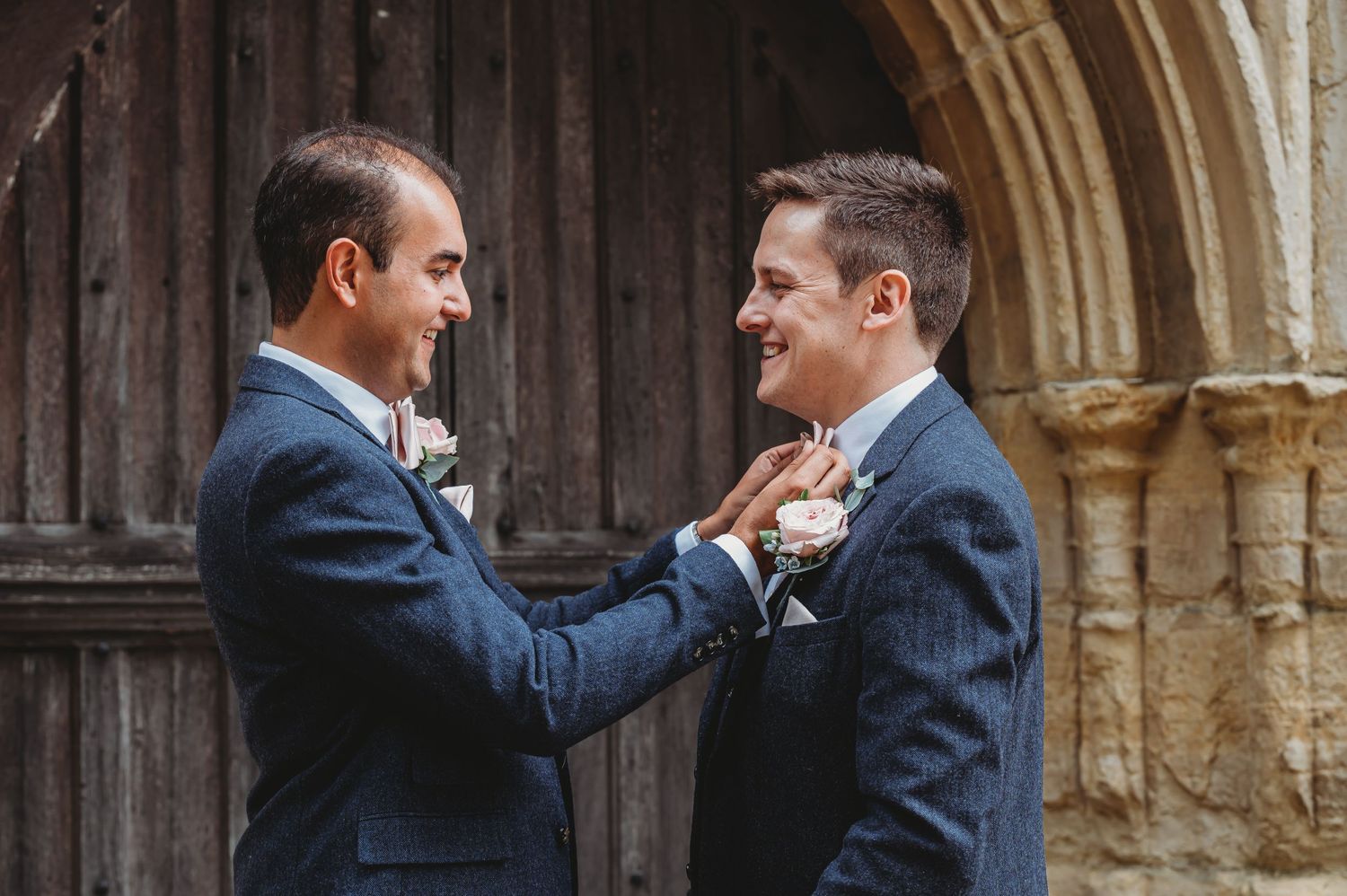 Two people in navy blue suits adjust each other's boutonnieres outside a stone archway during a wedding ceremony.