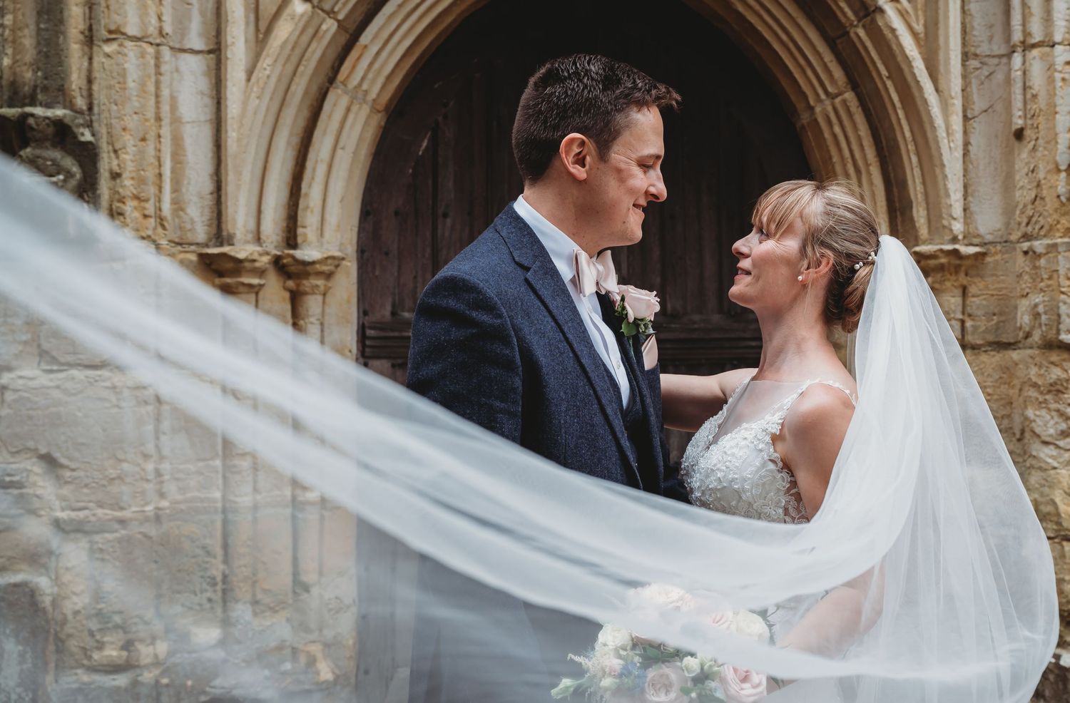 A romantic wedding moment captured at a gothic stone archway as the bride's veil flows in the breeze.