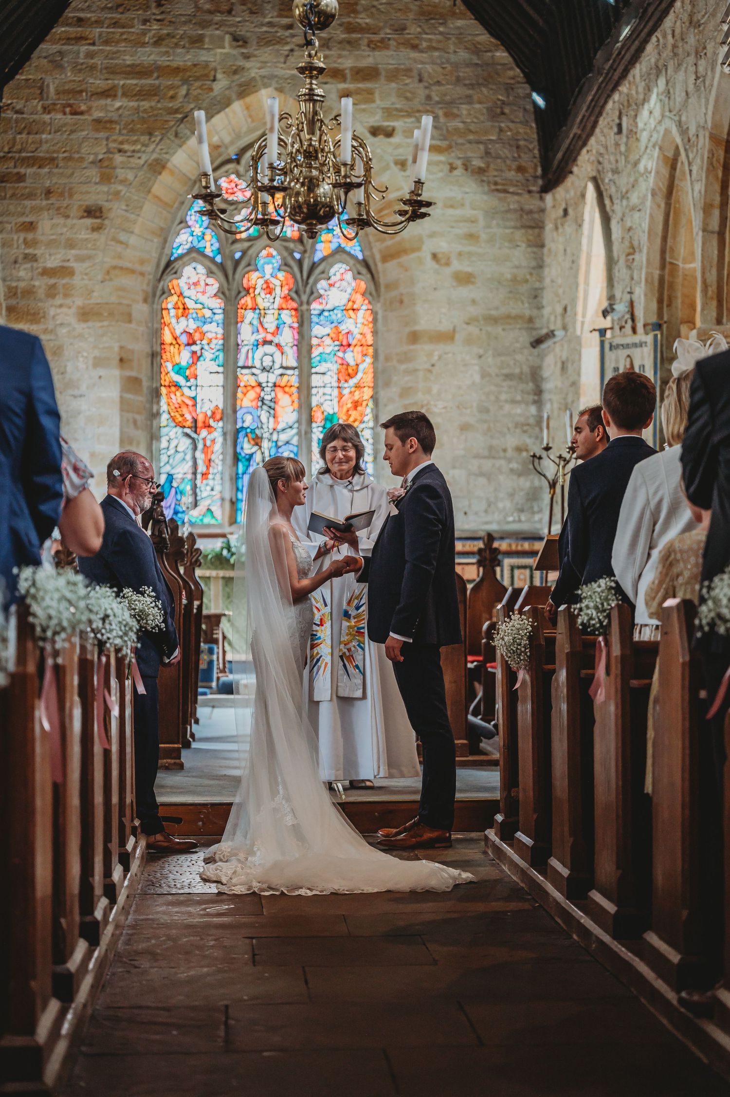 Wedding ceremony inside historic stone church with stained glass windows, antique chandelier, and floral pew decorations.