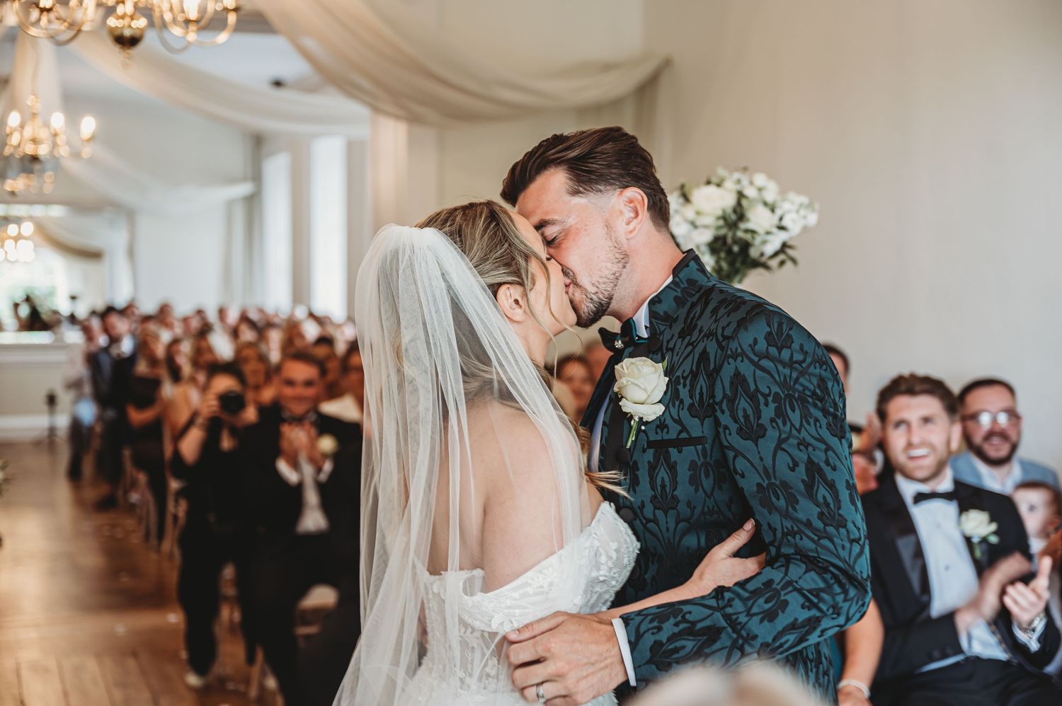 Wedding couple shares intimate first kiss in elegant white venue with guests looking on.