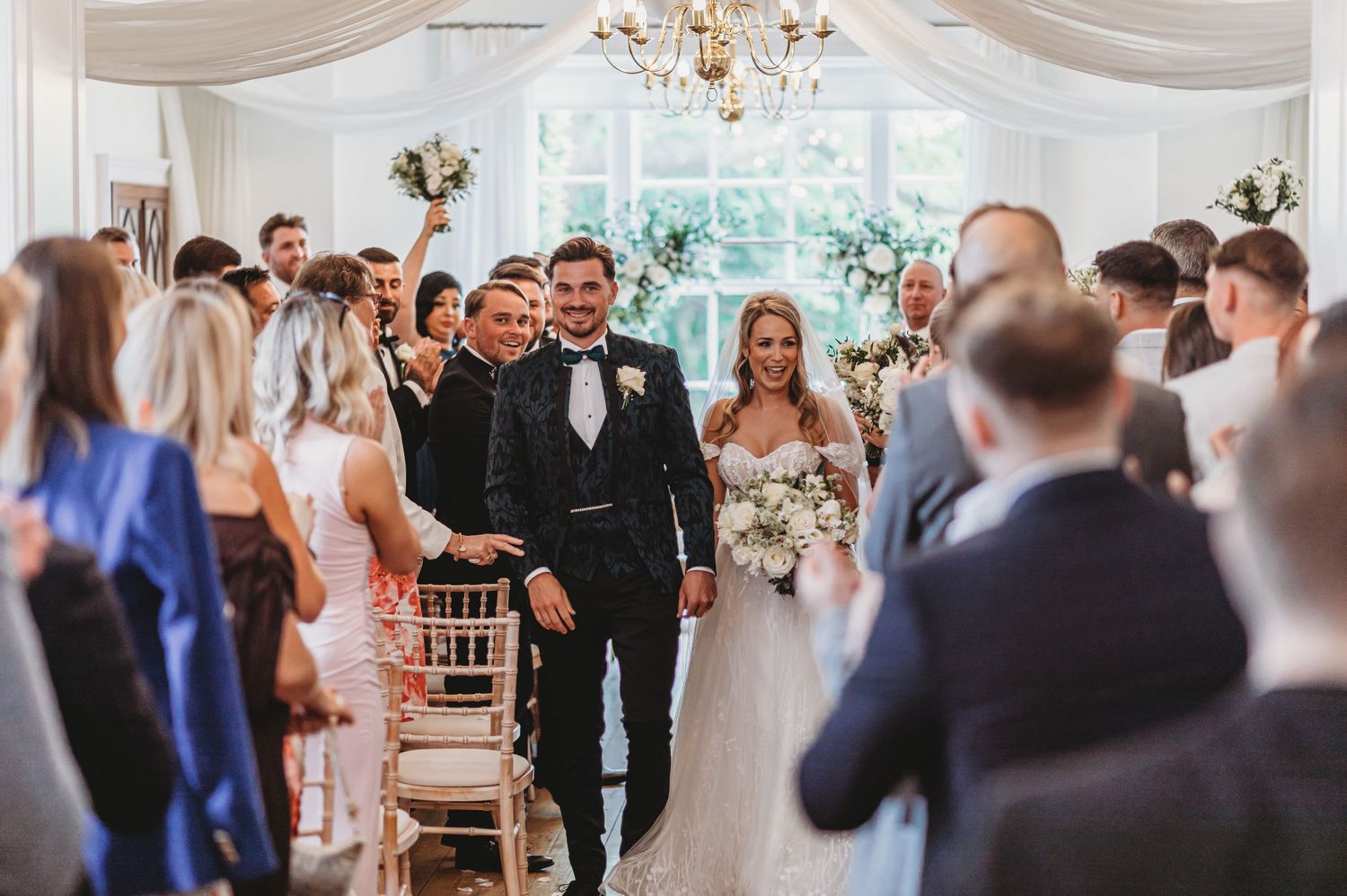 Wedding ceremony in an elegant white room with chandelier and gathered guests lining the aisle.