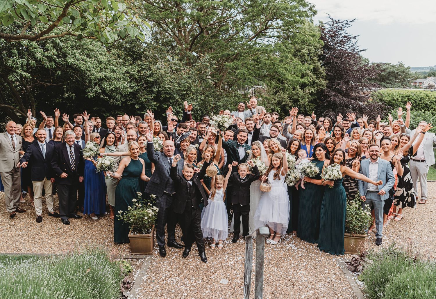Joyful wedding party and guests pose for large group photograph in garden setting.
