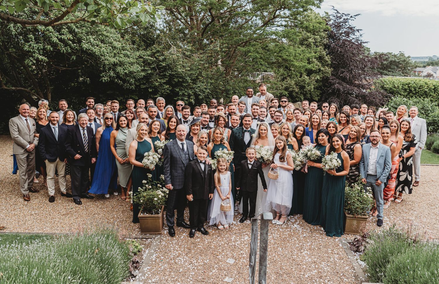 Large wedding group photo of guests and wedding party gathered outdoors on gravel path.
