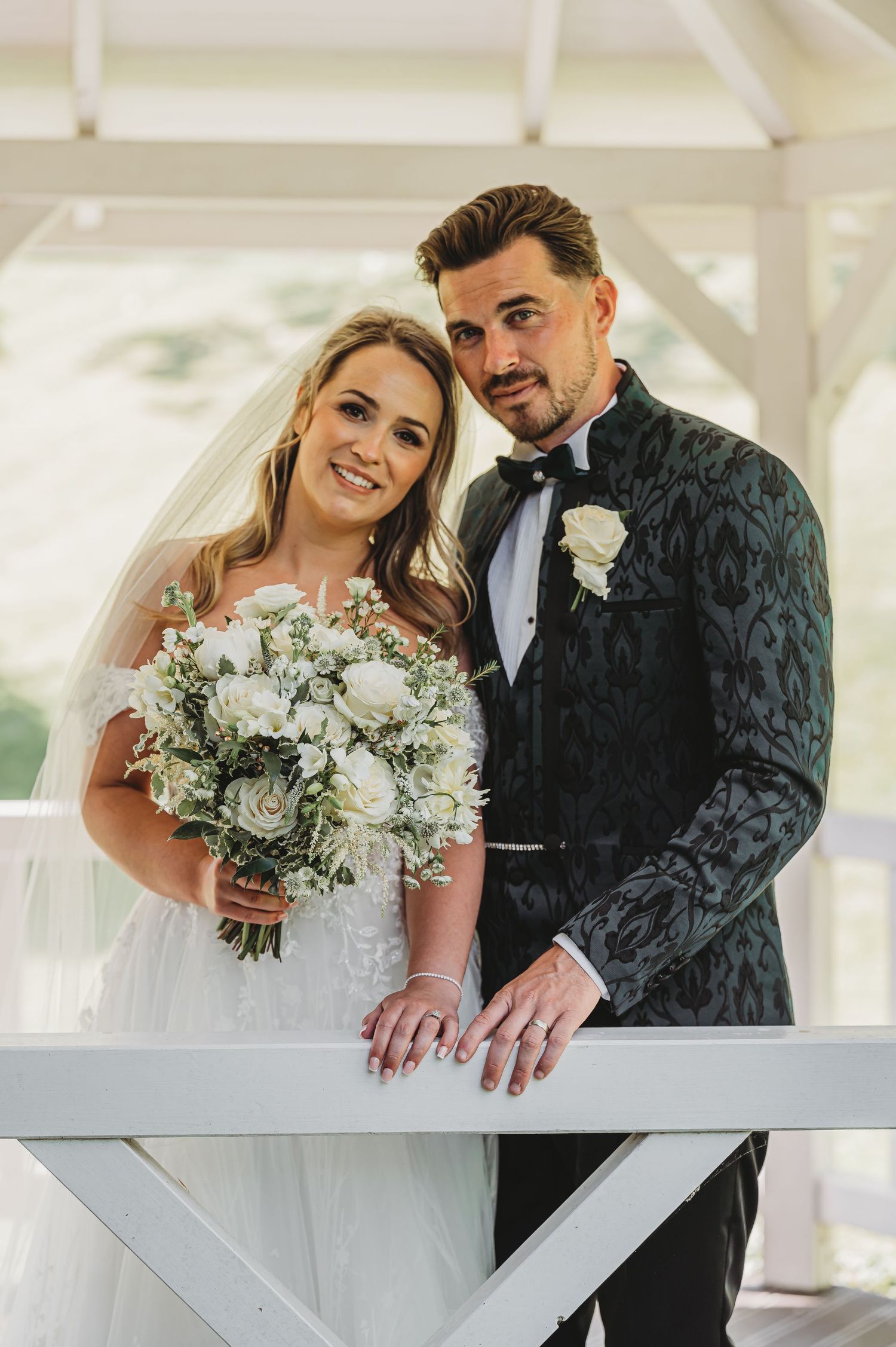 Elegant wedding portrait with bride holding lush white bouquet and groom in textured black jacket.