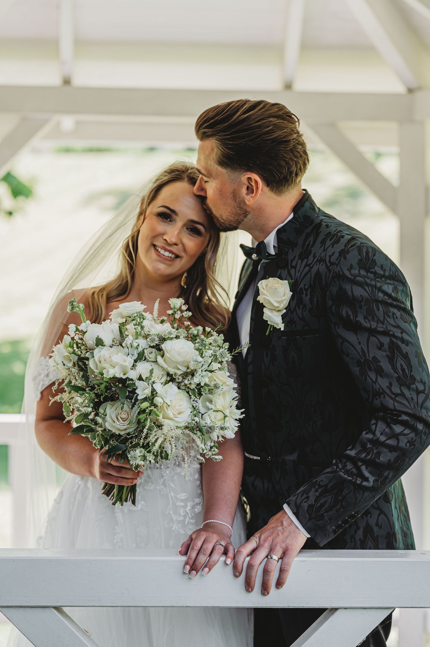 Wedding couple shares intimate moment with bride holding white and green bouquet against white backdrop.