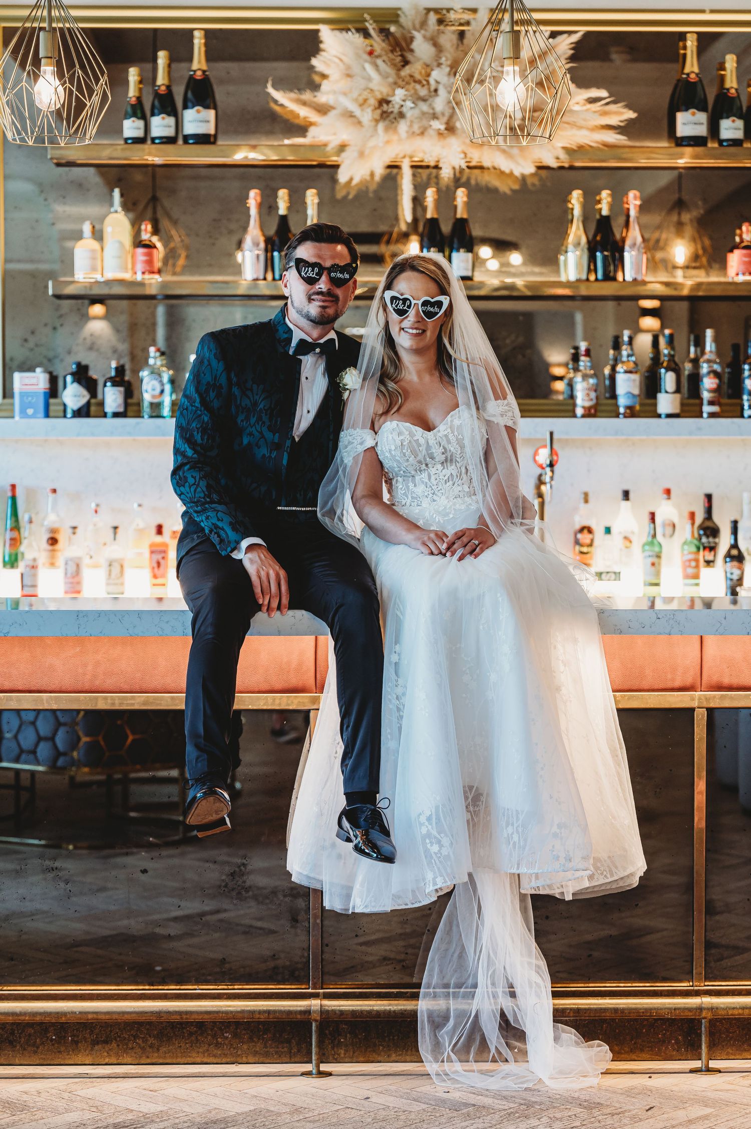 Stylish newlyweds pose at a bar wearing sunglasses with bottles and decor in background.