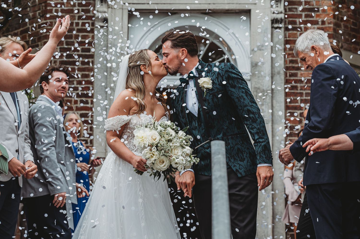 Newlyweds share a moment under brick archway while guests shower them with confetti.