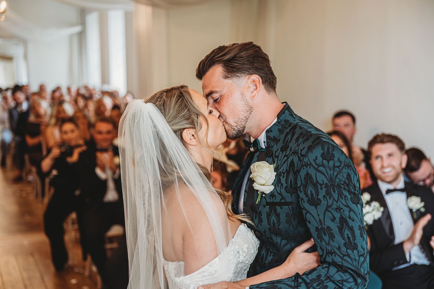 Romantic wedding moment captured in elegant white venue with guests seated in background.