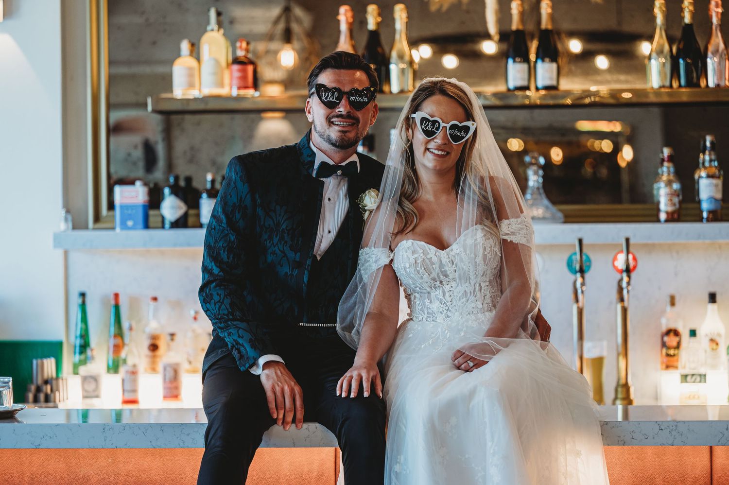 Newlyweds wearing sunglasses pose at reception bar area.