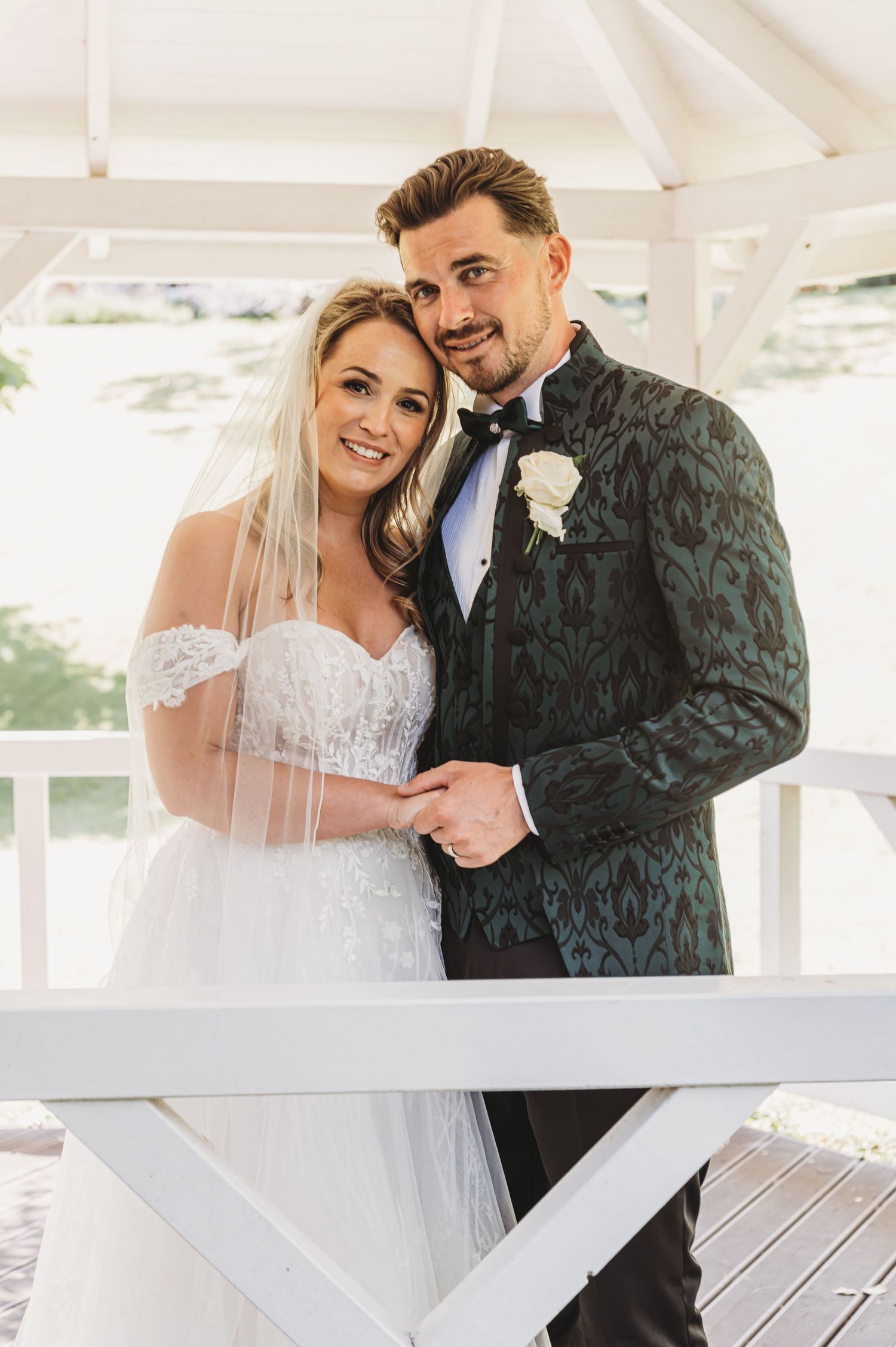Wedding couple poses in white gazebo with bride in lace gown and groom in patterned tuxedo.