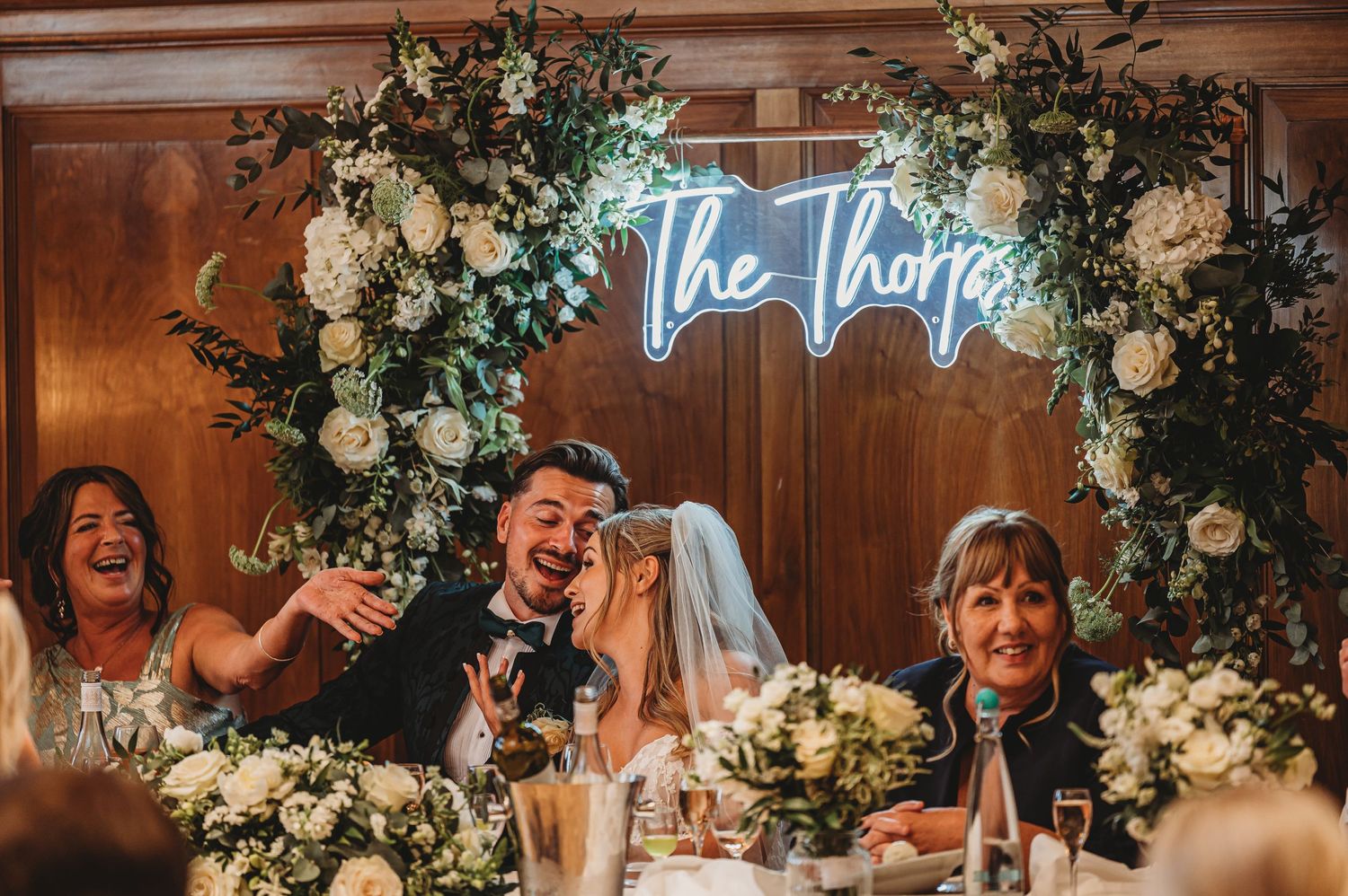 Wedding reception decor features neon sign reading 'The Thorpes' with white floral arrangements.