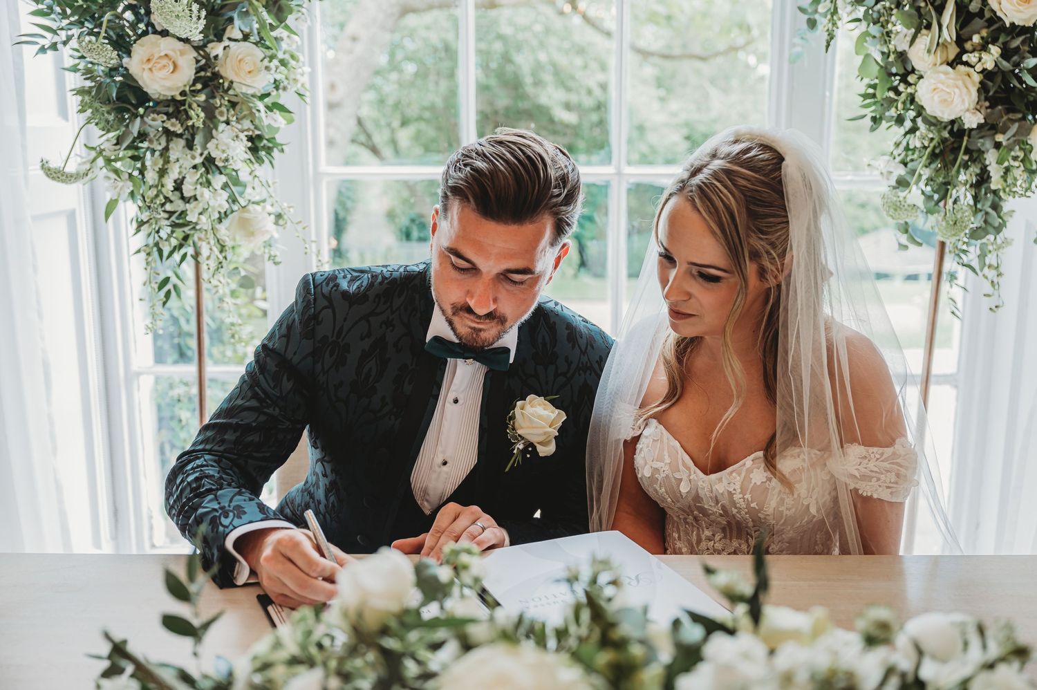 Couple signing marriage documents at elegant wedding venue decorated with white floral arrangements.