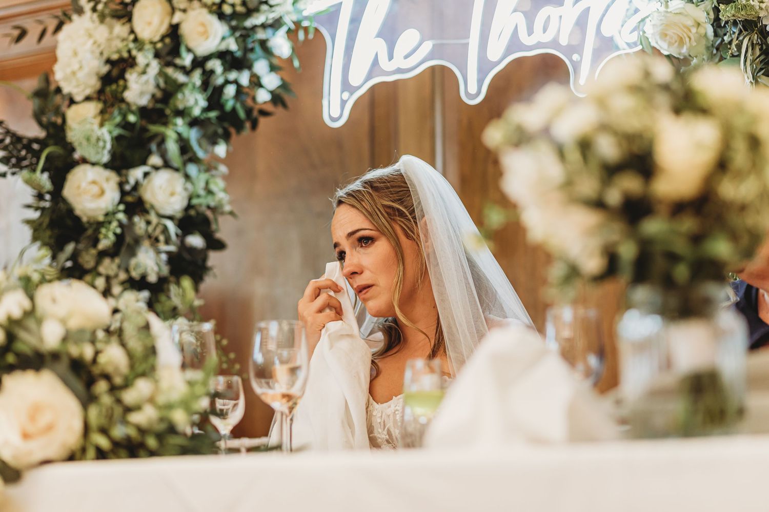 A bride in white veil sits at decorated reception table with elegant floral arrangements.